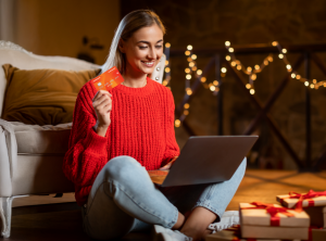 Mujer con gorro navideño mirando el computador y sosteniendo su tarjeta Cencosud para comprar regalos para Navidad Tarjeta Cencosud.