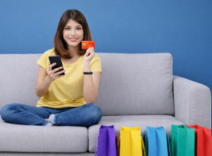 Joven sentada entre bolsas de regalo usando su teléfono móvil y una tarjeta bancaria para reservar viajes en semana santa 2026